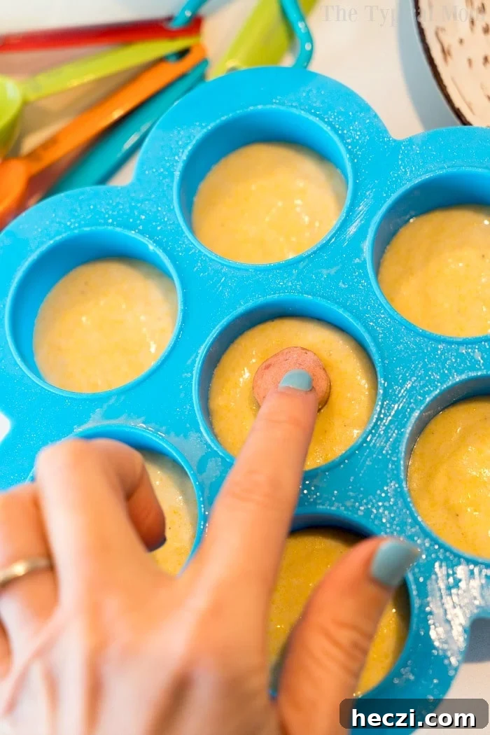 Pressure cooker corn dog bites are easy to make in your Instant Pot, Crock Pot Express or Ninja Foodi! A hand places a mini hot dog into cornbread batter in a blue silicone mold, preparing pressure cooker corn dog bites. Colorful measuring spoons are nearby.