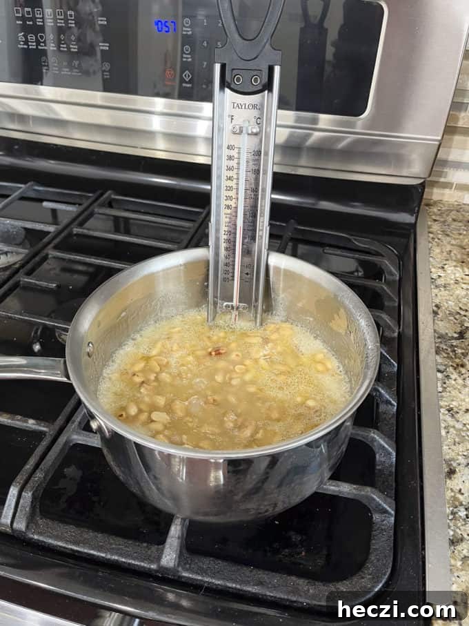 Classic Peanut Brittle Recipe: Crispy, Homemade Candy in 30 Minutes 11 a pot on a stovetop with granulated sugar, water, corn syrup and peanuts cooking in it and a candy thermometer attached to the side