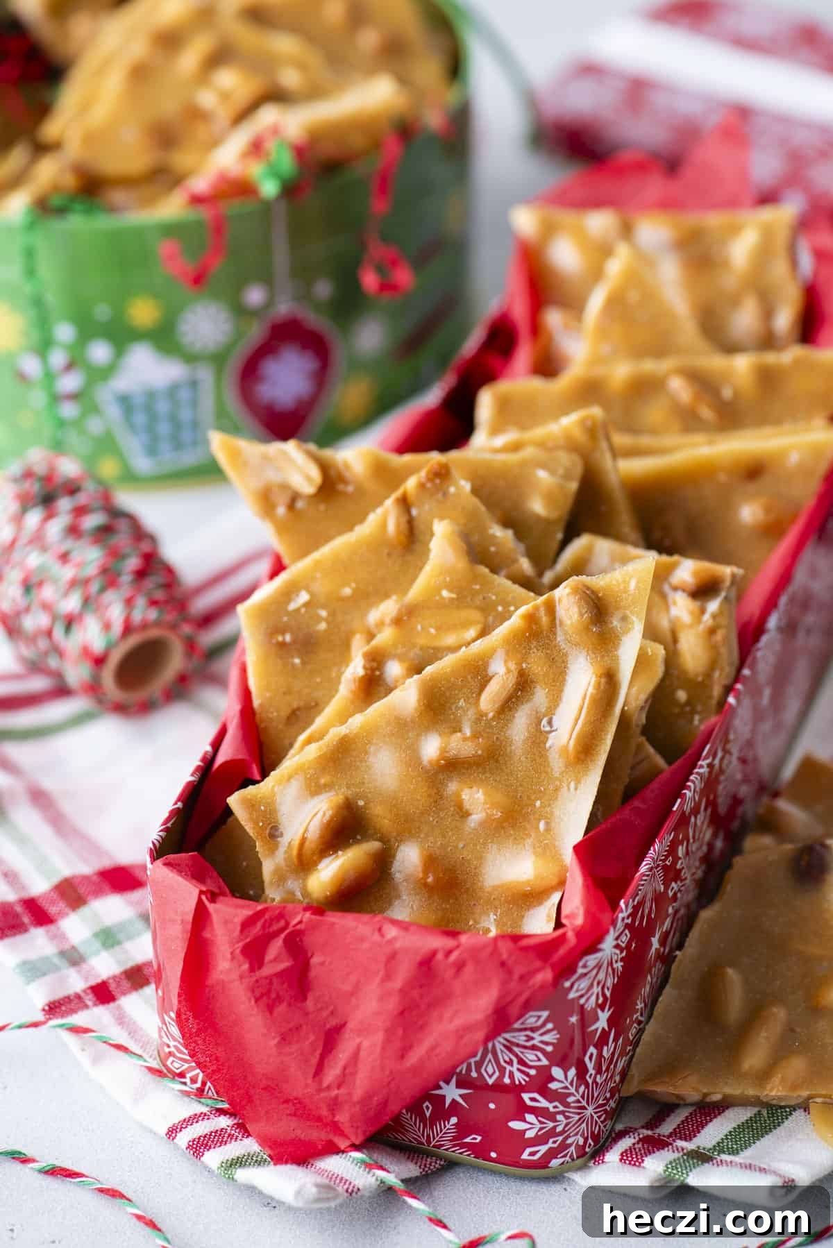 Classic Peanut Brittle Recipe: Crispy, Homemade Candy in 30 Minutes 4 a red christmas tin full of peanut brittle with a green christmas tin in the background, red, white and green string, all sitting on top of a red, green and white kitchen towel.