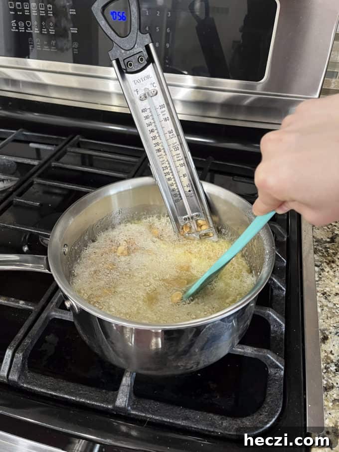Classic Peanut Brittle Recipe: Crispy, Homemade Candy in 30 Minutes 10 a pot on a stovetop with granulated sugar, water, corn syrup and peanuts cooking in it being mixed with a rubber spatula and a candy thermometer attached to the side