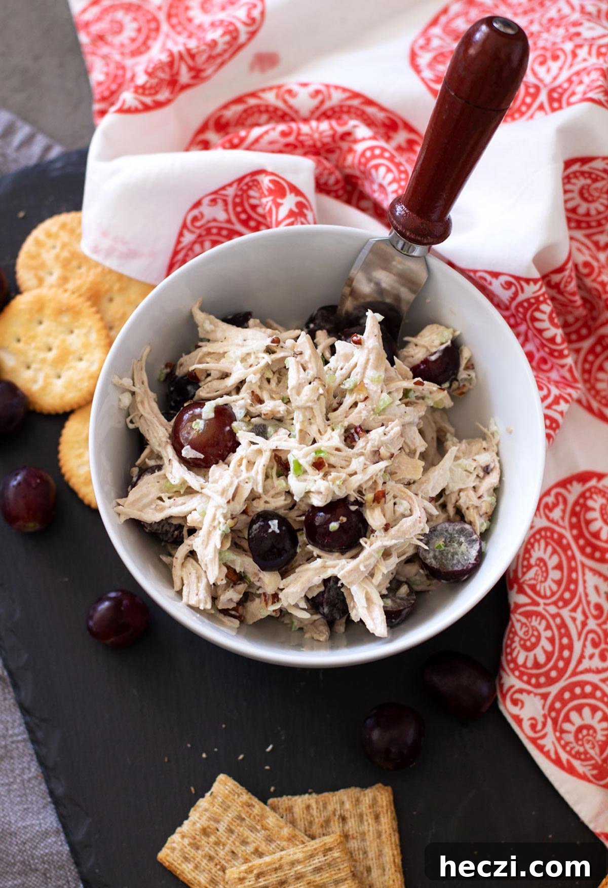 White bowl with a serving knife containing chicken salad topped with red grapes, crackers on table.