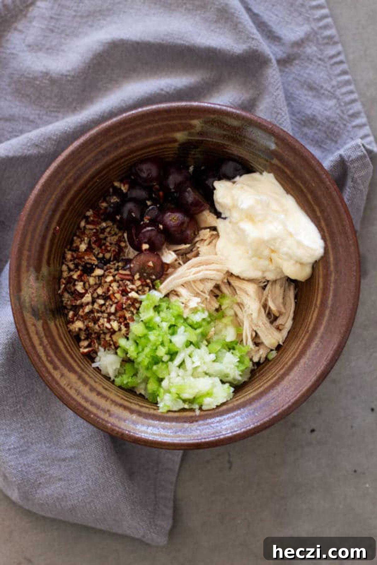 Brown bowl containing pecan chips, chopped celery, onion, red grapes, mayonnaise and shredded chicken.