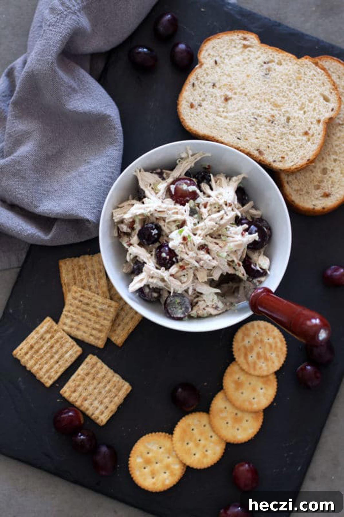 White bowl of chicken salad on a black serving tray, bread, crackers and grapes on serving dish.