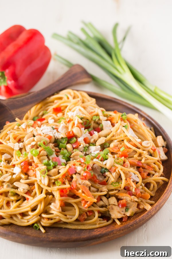Easy Spicy Peanut Noodles With Chicken on a wood serving plate with a wood serving spoon, and a whole red pepper and a bundle of green onions in the background. 