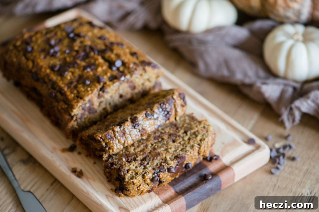 Sliced Pumpkin Oatmeal bread with chocolate chips laying on a wooden cutting board. The light colored wood table is adorned with white and orange pumpkins and a light brown piece of fabric. A small knife sits beside the board. 