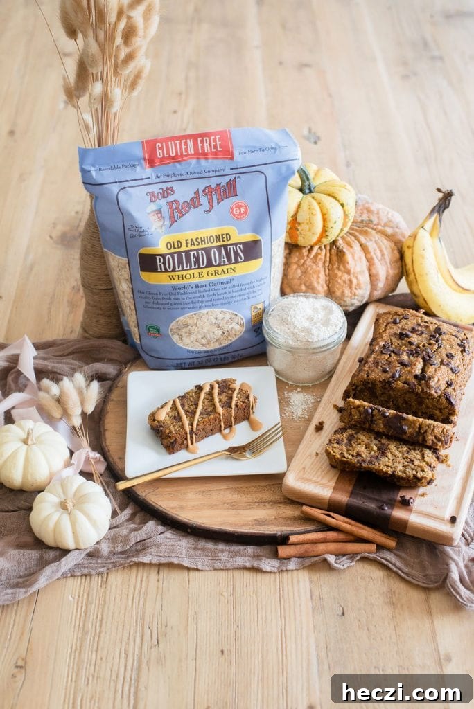 A bag of Bob's Red Mill Old Fashioned Gluten Free Rolled Oats sits in the middle of a festive Autumn table with white, yellow, and orange pumpkins and decorative wheat. A piece of banana bread and peanut butter is on a white plate and a wooden cutting board sits nearby by the full loaf of bread. 