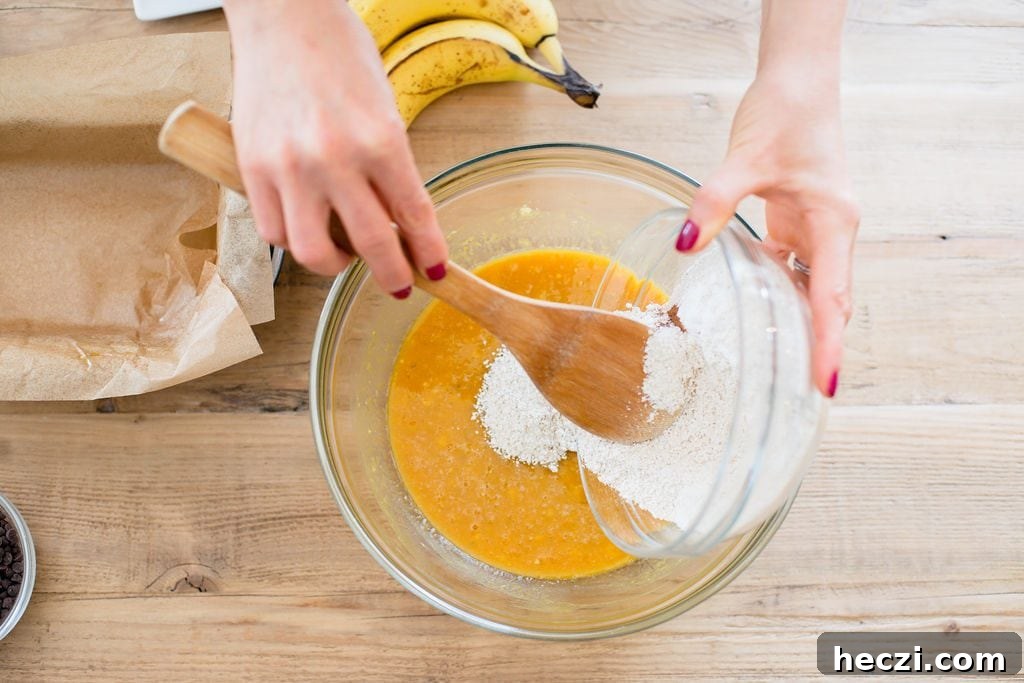 Mixing dry ingredients for Banana Pumpkin Oatmeal Bread into the wet ingredients. One hand is holding the small glass bowl and the other hand is stirring with a wooden spoon. Bananas are laying on the table, as well as a bowl of chocolate chips and a loaf pan with parchment paper. 
