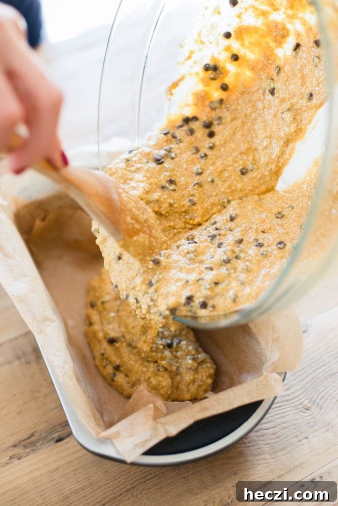 A woman's hand is shown using a wooden spoon to pour oatmeal banana pumpkin bread into a loaf pan lined with parchment paper. 