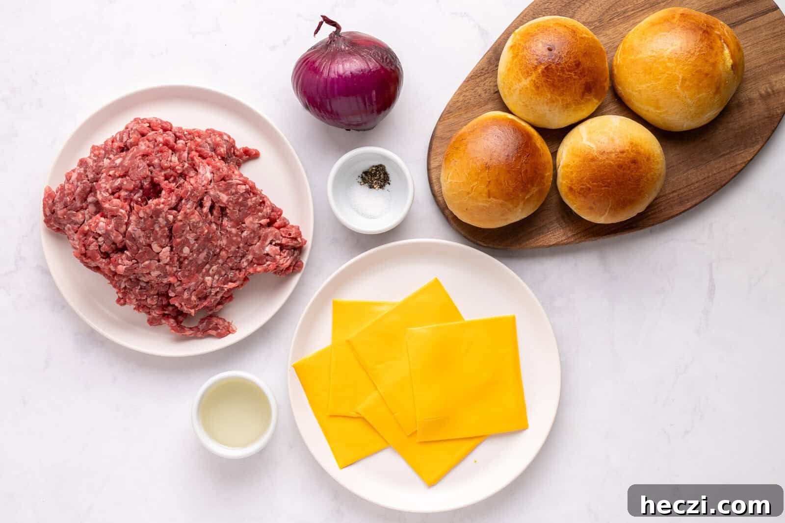 How to Make a Crispy Smash Burger at Home 3 Top-down view of burger ingredients: a plate of ground beef, a red onion, salt and pepper, four burger buns on a wooden board, American cheese slices on a plate, and a small bowl of oil on a white surface.