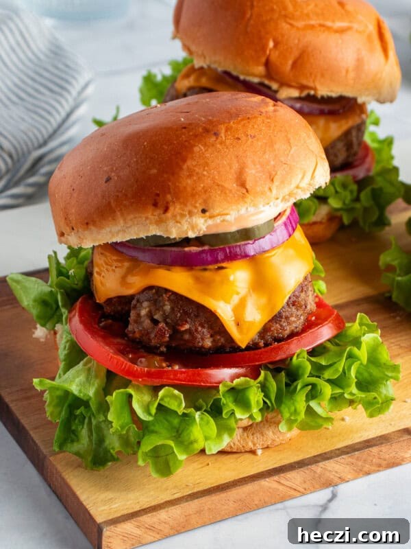 How to Make a Crispy Smash Burger at Home 5 Two cheeseburgers on a wooden board with lettuce, tomato, cheddar cheese, red onion, and pickles, on a brioche bun. A striped napkin is in the background.