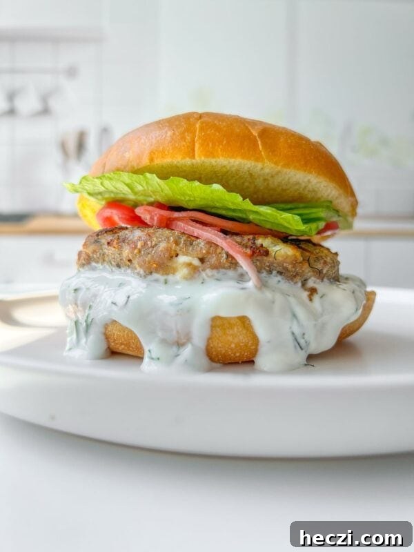 How to Make a Crispy Smash Burger at Home 6 A close-up of a burger on a white plate. The burger features a creamy white sauce dripping down, a patty, tomato slices, and fresh lettuce, all sandwiched in a toasted bun. The background is a blurred kitchen setting.