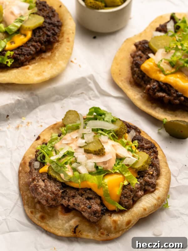 How to Make a Crispy Smash Burger at Home 7 Three open-faced cheeseburger flatbreads topped with melted cheese, lettuce, chopped onions, sliced pickles, and sauce, on parchment paper. A small bowl of extra pickles is in the background.