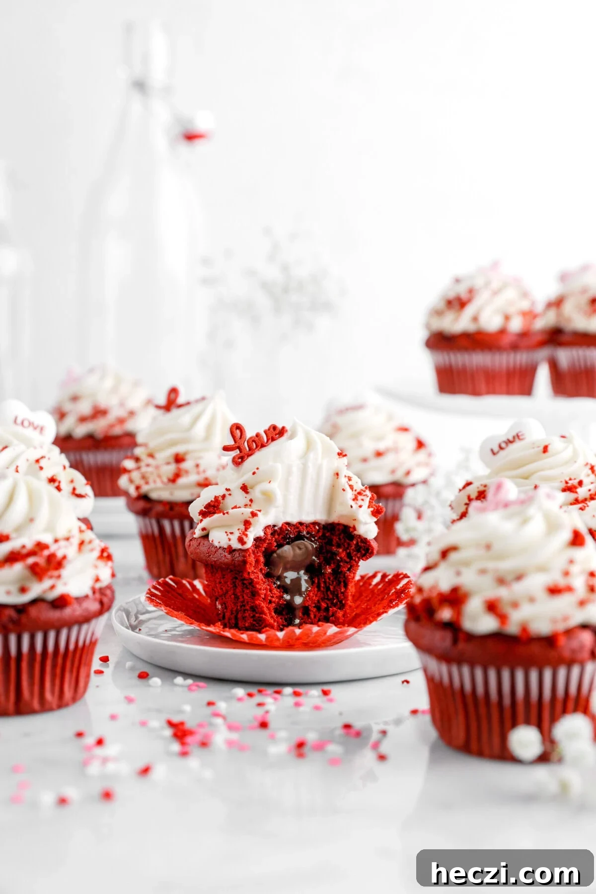 Red Velvet Molten Lava Cupcakes: Gooey Chocolate Center Delight 3 A red velvet cupcake with cream cheese frosting sits on a white plate, its wrapper peeled back to reveal a molten chocolate center. A red "love" topper decorates the swirl of frosting, with red velvet crumbs sprinkled on top. Surrounding cupcakes, heart-shaped sprinkles, and soft-focus white flowers create a romantic, festive scene.