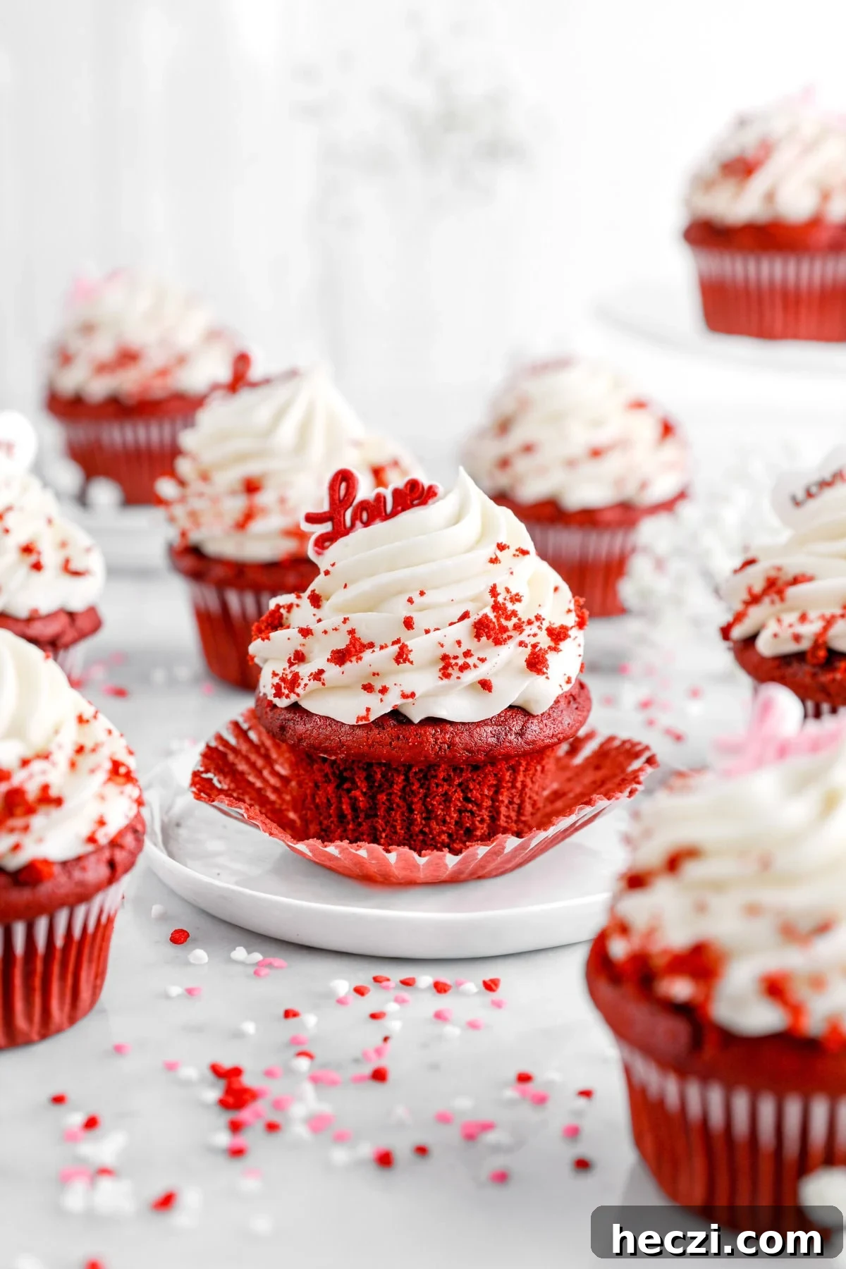Red Velvet Molten Lava Cupcakes: Gooey Chocolate Center Delight 4 A red velvet cupcake with cream cheese frosting sits on a white plate, its wrapper partially peeled back to reveal the moist cake. Red velvet crumbs and festive heart-shaped sprinkles are scattered around, while more cupcakes with swirls of frosting and "love" toppers decorate the blurred background.