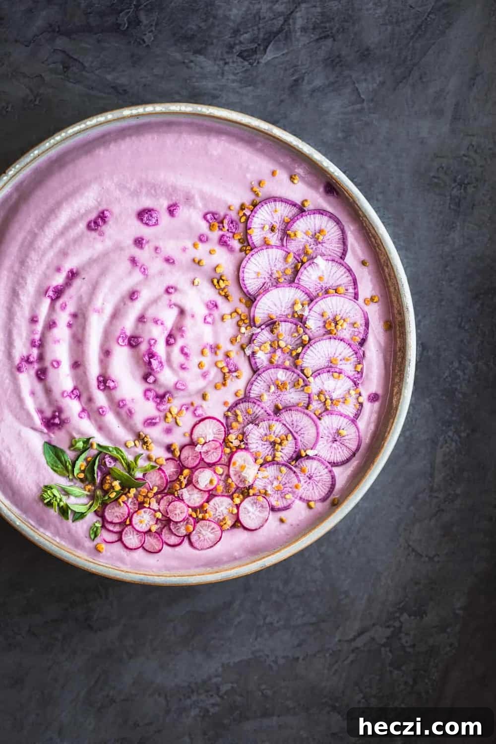 Vegan Purple Cauliflower Soup with Sliced Purple and Pink Radishes, tiny basil leaves, Bee Pollen and Flaky Sea Salt. Overhead shot, left side of bowl is cut off, on a dark grey background.