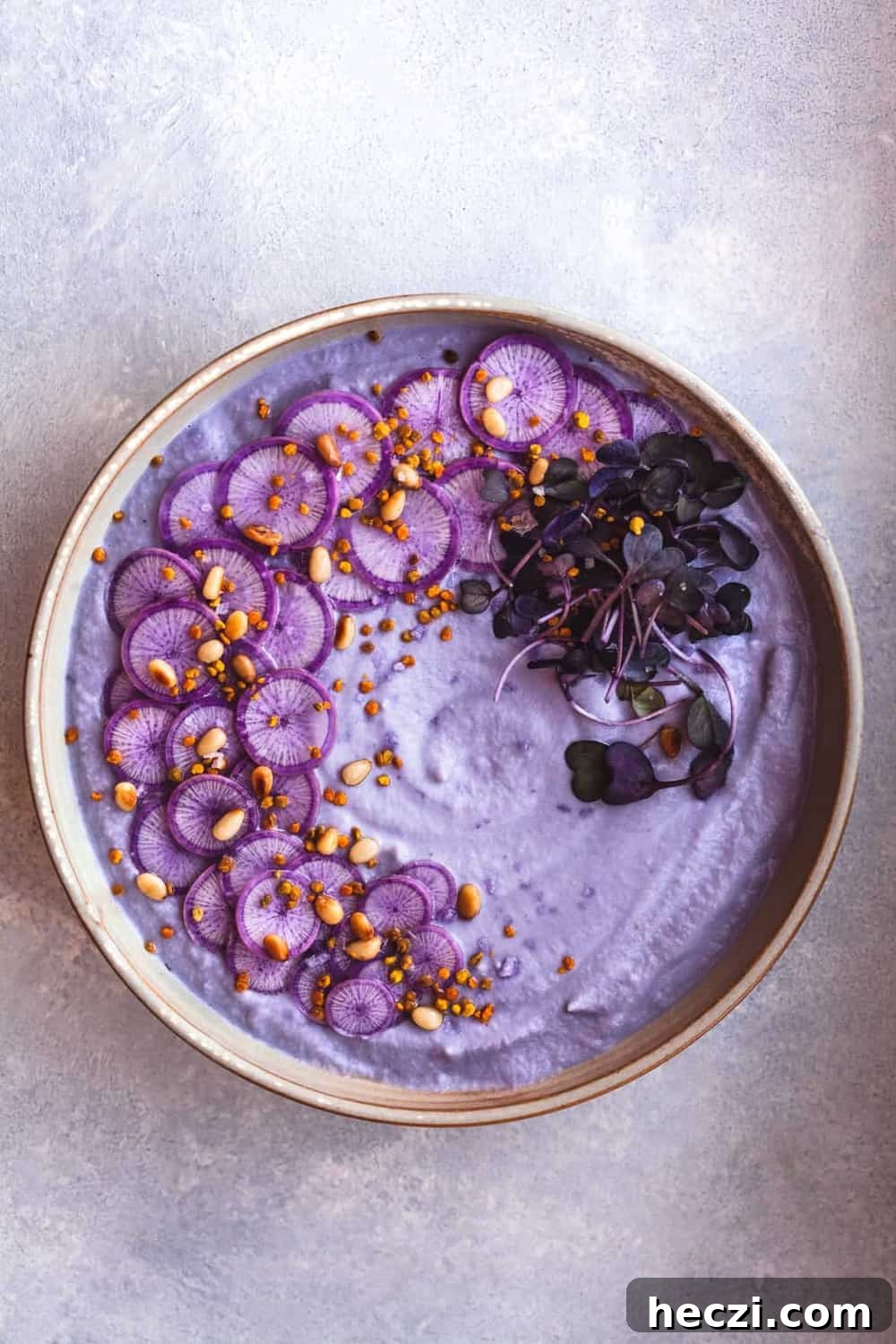 Vegan Cauliflower Soup with Purple Ninja Radishes, Daikon Radish Sprouts, Pine Nits & Bee Pollen. Overhead shot. Soup Bowl is in center of the frame on a white/light blue background.