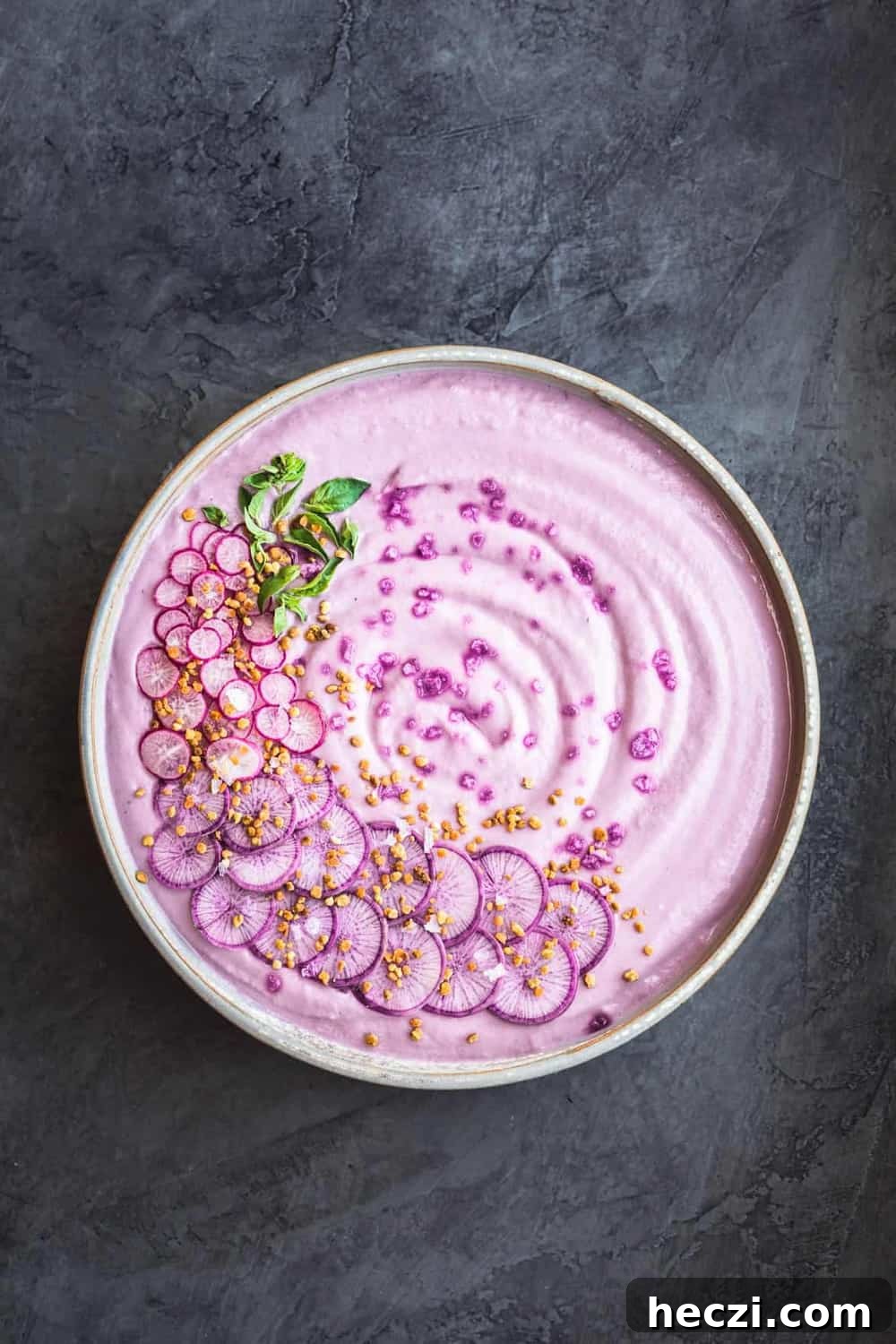 Vegan Purple Cauliflower Soup with Sliced Purple and Pink Radishes, tiny basil leaves, Bee Pollen and Flaky Sea Salt. Overhead shot. Bowl is in center of the frame on a dark grey background.
