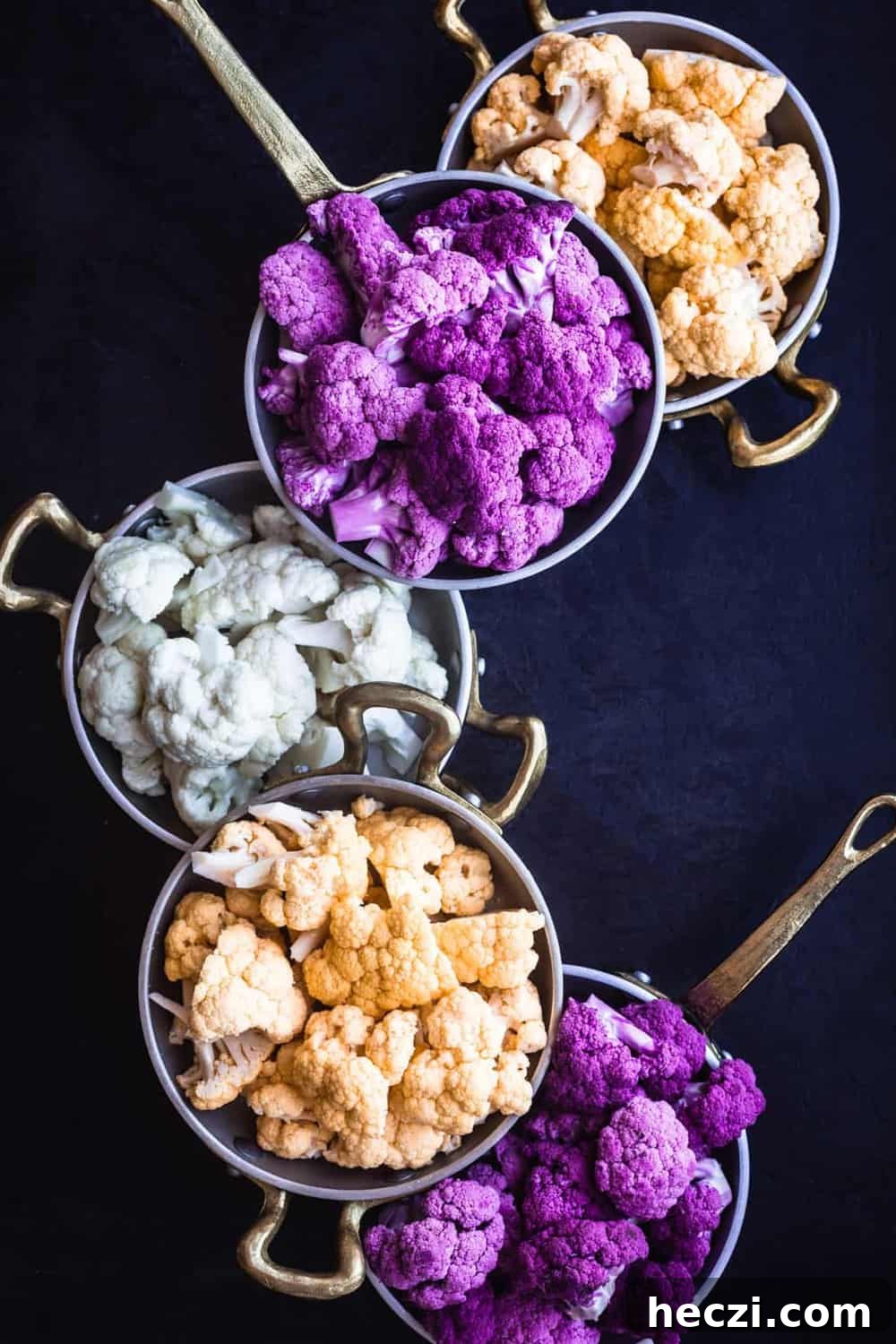 Purple, Yellow/Orange and White Cauliflower cut into florets and separated by color, into 5 little pans (2 of purple, 2 of yellow/orange and 1 white) on a black background.