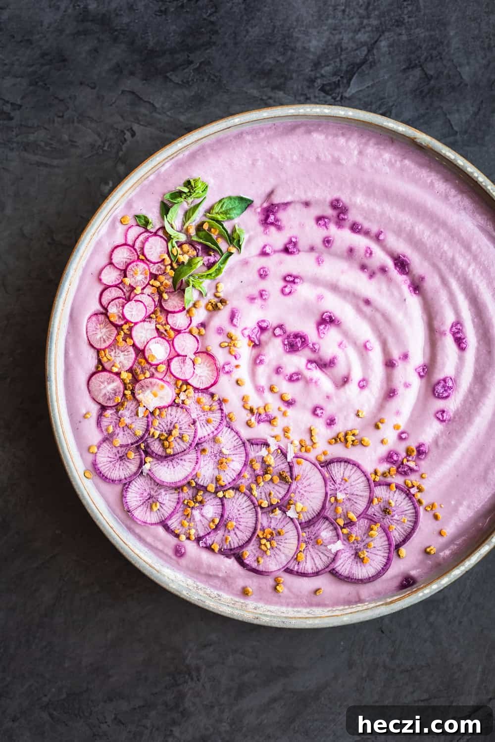 Vegan Purple Cauliflower Soup with Sliced Purple and Pink Radishes, tiny basil leaves, Bee Pollen and Flaky Sea Salt. Overhead shot, right side of bowl is cut off, on a dark grey background.