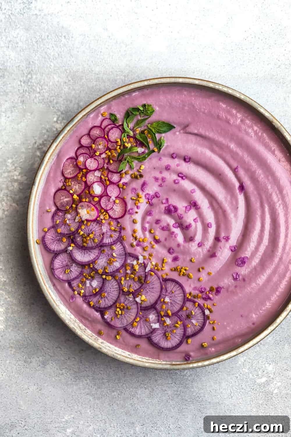 VeganPurple Cauliflower Soup with Sliced Purple and Pink Radishes, tiny basil leaves, Bee Pollen and Flaky Sea Salt. Overhead shot, right side cut off, on a white/light blue background.
