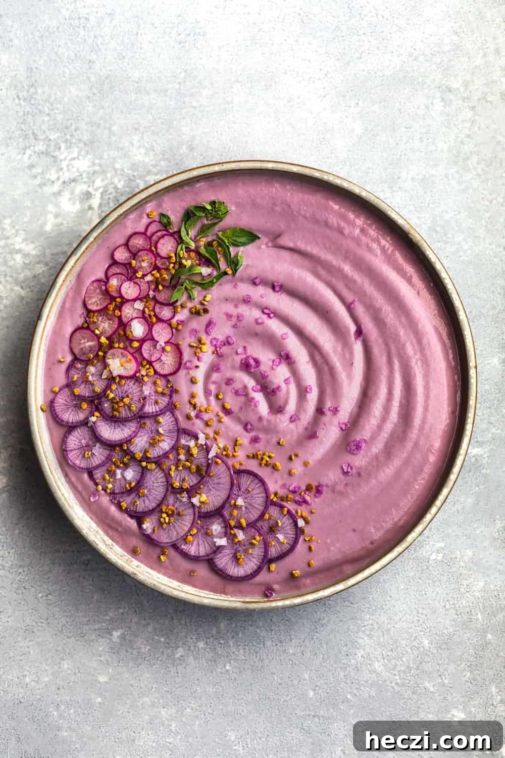 Vegan Purple Cauliflower Soup with Sliced Purple and Pink Radishes, tiny basil leaves, Bee Pollen and Flaky Sea Salt. Overhead shot. Bowl is in center of the frame on a white/light blue background.