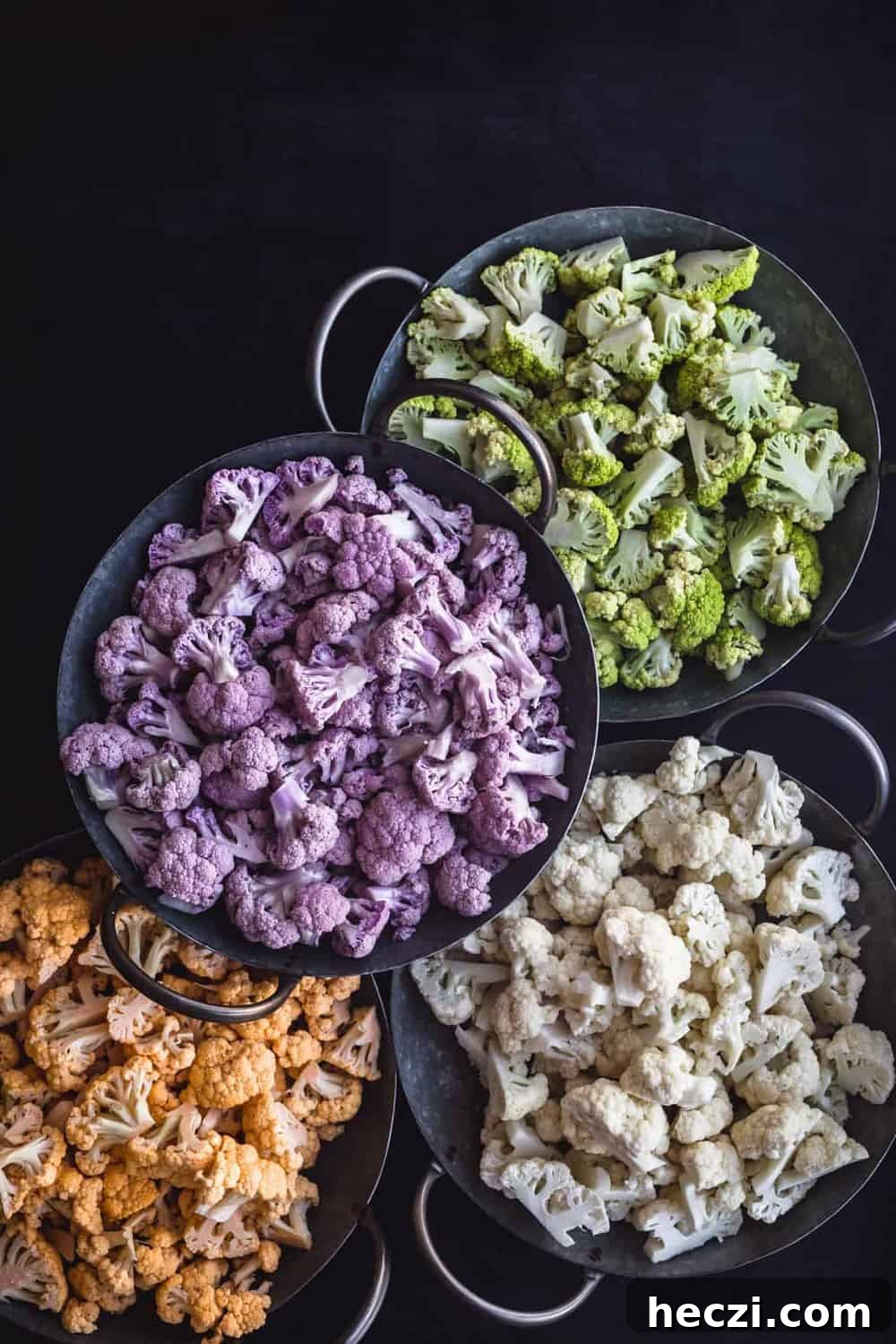 Yellow/Orange, Purple, White, and Green Cauliflower, each cut into florets and separated by color into 4 silvers bowls with handles, on a black background.