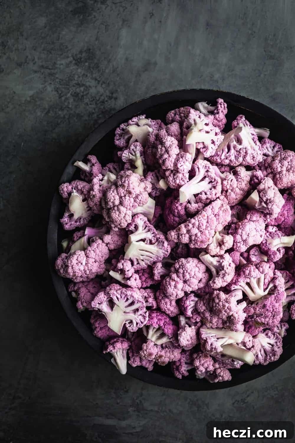 Bowl of purple cauliflower cut into florets. Overhead shot, right side cut off, on a dark grey background.