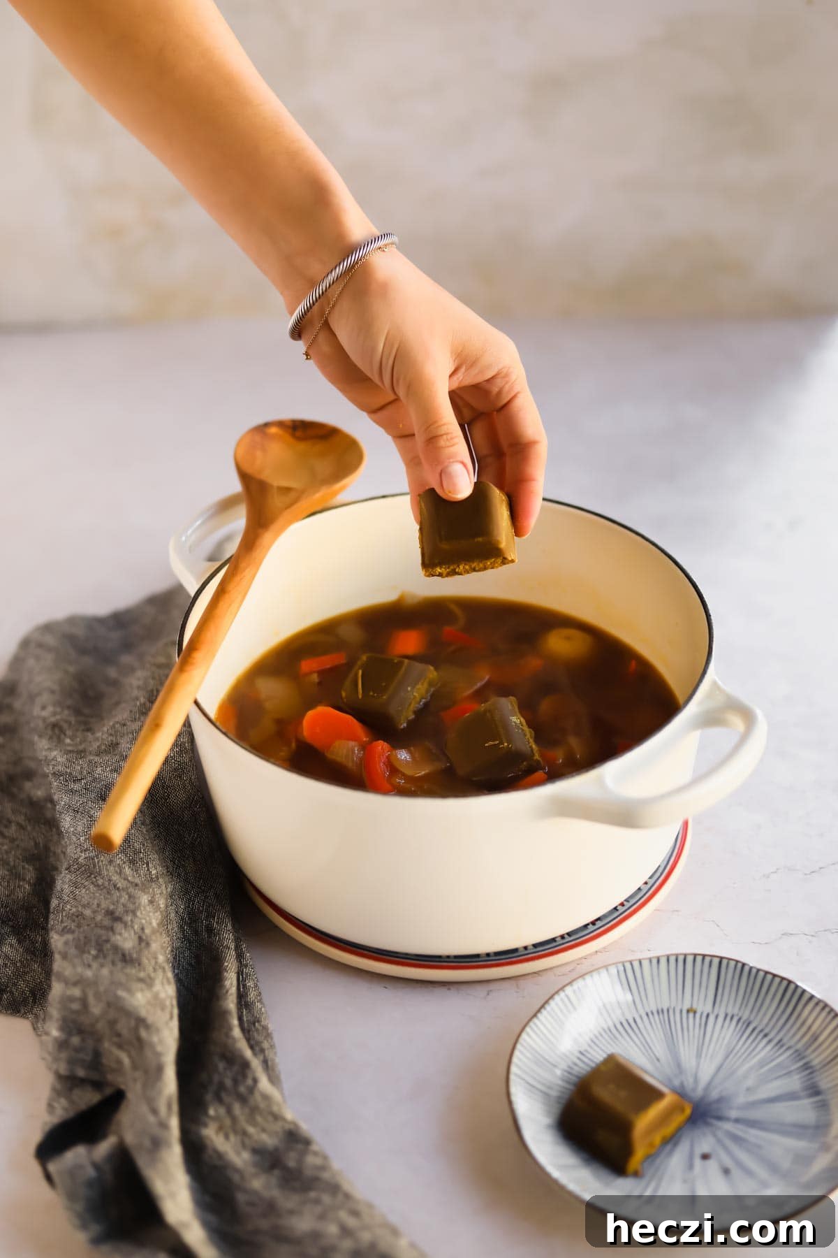 Japanese Chicken Katsu Curry Recipe — Crispy Cutlet with Rich Curry 7 Placing Golden Curry Rou in white Le Creuset dutch oven containing on onion, potatoes, and bell peppers in with wooden spoon