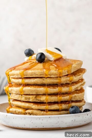 Syrup being poured over a stack of oat flour pancakes topped with blueberries.