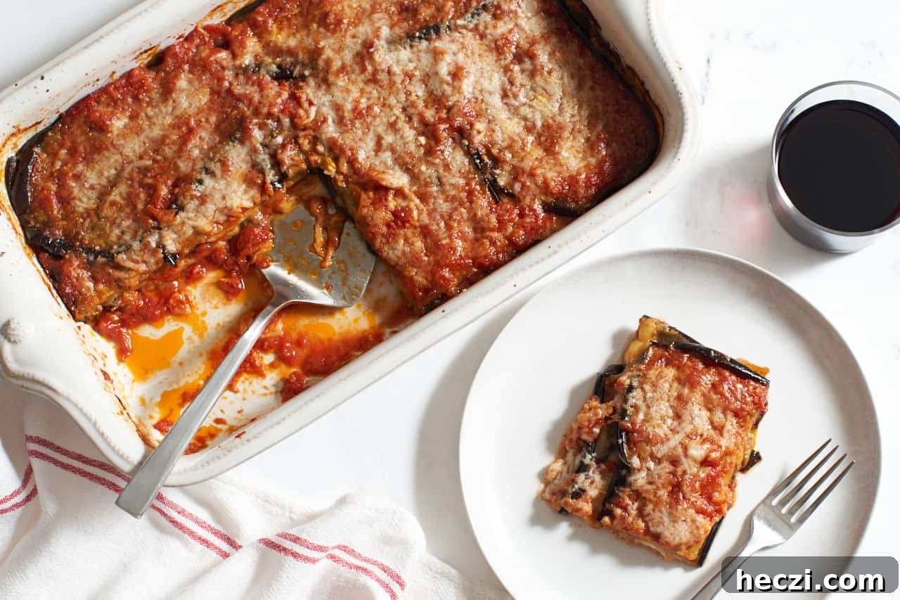 A casserole dish of vegetarian eggplant lasagna with a spatula in it next to a glass of red wine, and a plate with a slice of lasagna on it.
