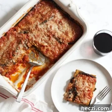 A casserole dish of vegetarian eggplant lasagna with a spatula in it next to a glass of red wine, and a plate with a slice of lasagna on it.