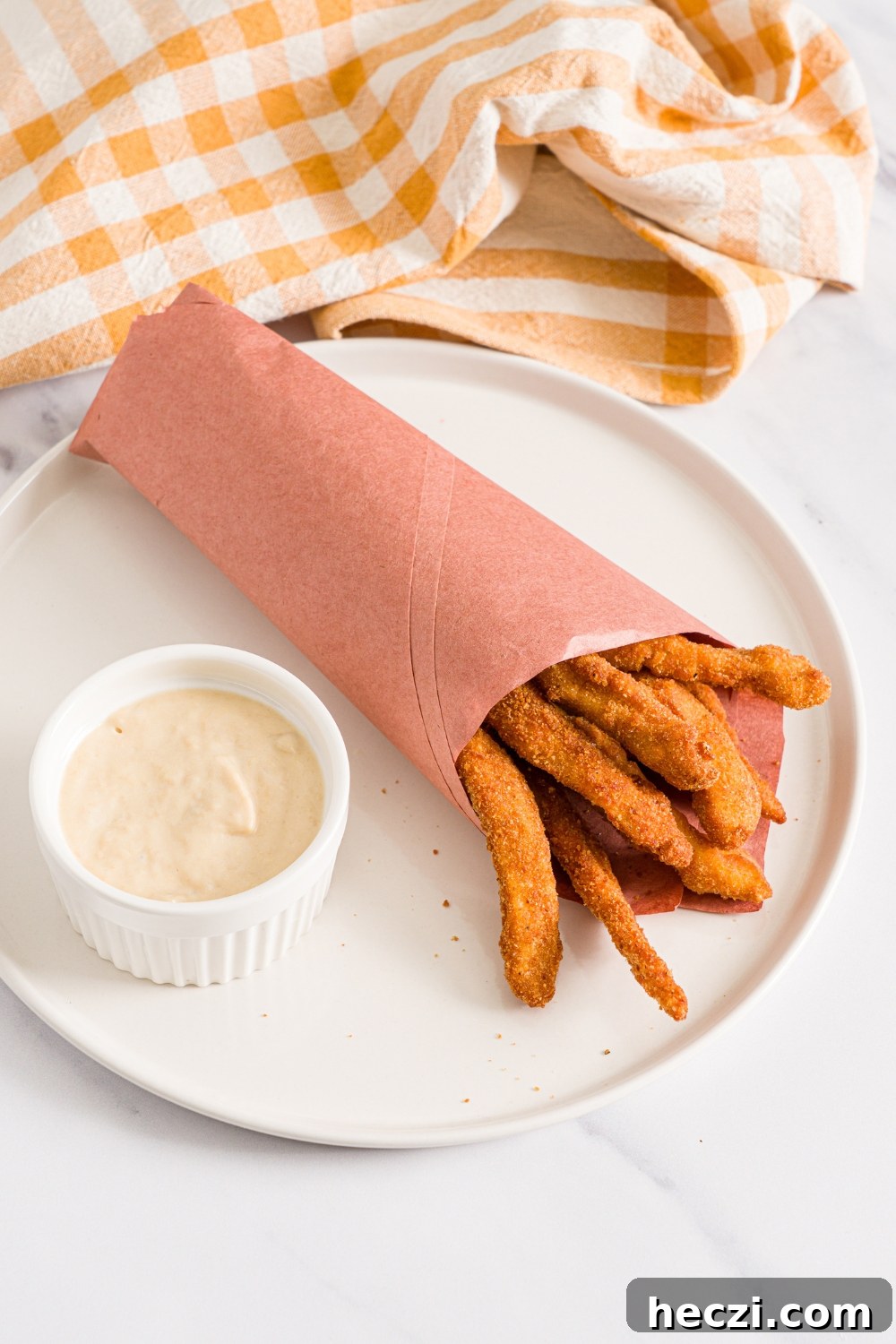 A white plate with fried homemade chicken fries wrapped in contact paper with a small ramekin of garlic aioli. The plate is on a marble counter with a yellow checkered napkin.