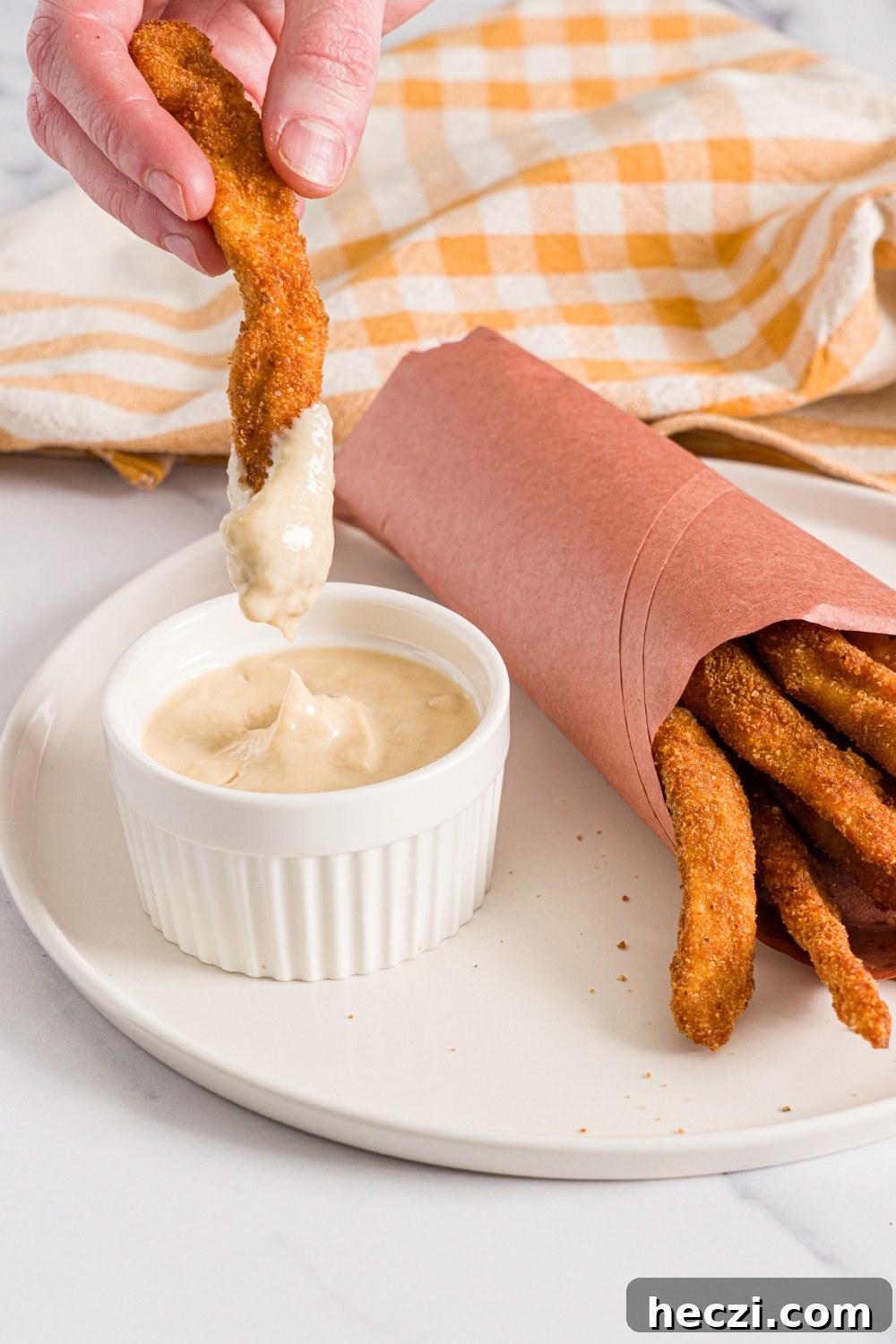 A white plate with fried homemade chicken fries wrapped in contact paper with a small ramekin of garlic aioli. A hand is dipping a chicken fry into the aioli.The plate is on a marble counter with a yellow checkered napkin.