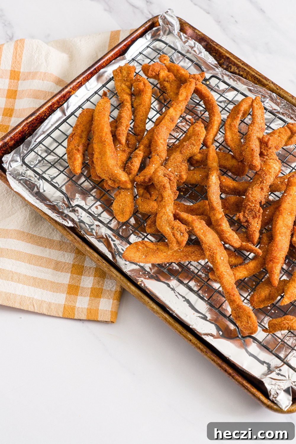 A baking sheet lined with foil and with a rack with fried chicken fries seasoned with salt. The sheet is on a marble counter with a yellow checkered napkin.