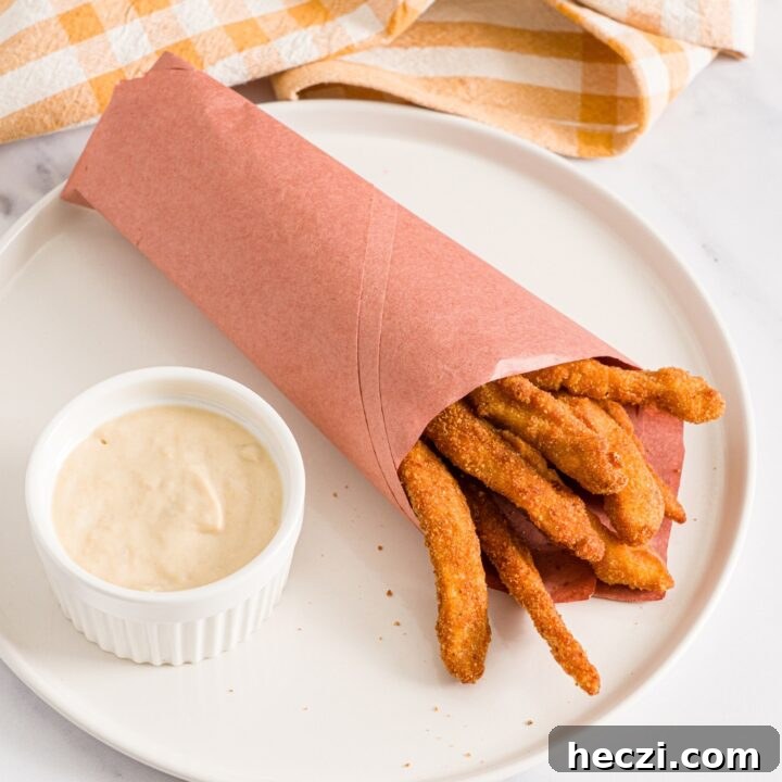 A white plate with fried homemade chicken fries wrapped in contact paper with a small ramekin of garlic aioli. The plate is on a marble counter with a yellow checkered napkin.
