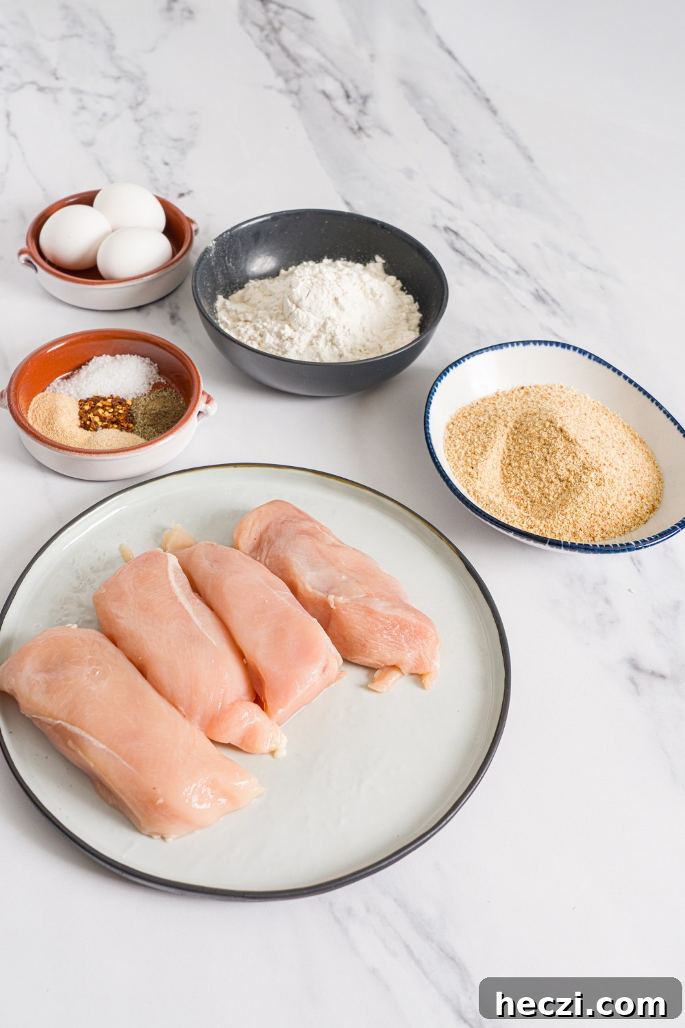 Several bowls in various sizes containing ingredients to make homemade chicken fries including chicken breasts, flour, breadcrumbs, eggs, and seasonings.