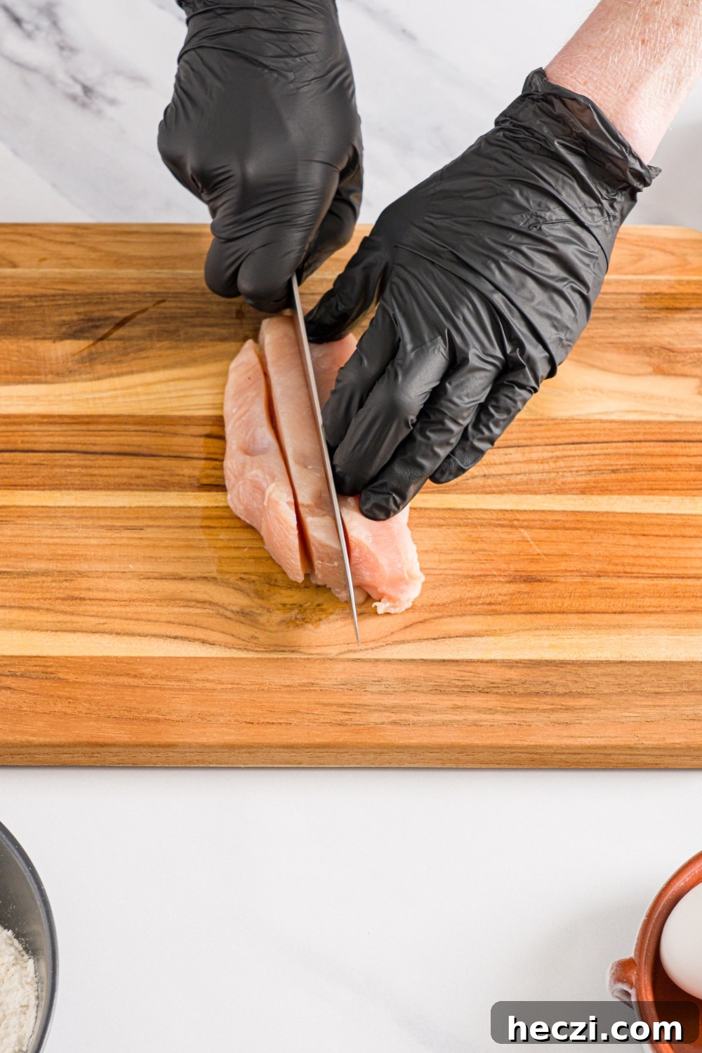 An uncooked chicken breast being sliced into strips on a wooden board. The board is on a marble counter.
