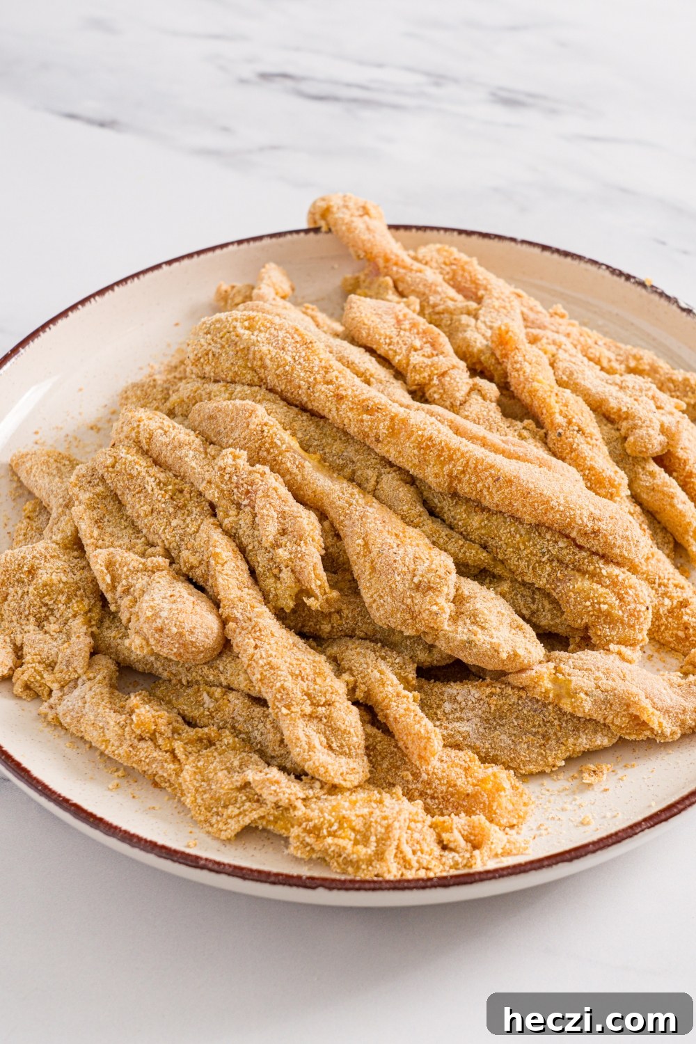 A plate with breaded uncooked chicken fries. The plate is on a marble counter.