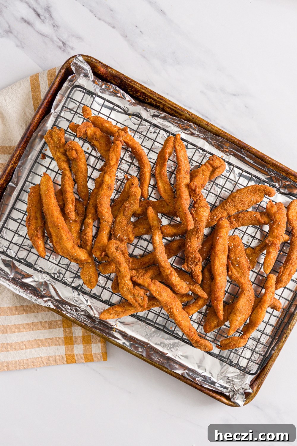 A baking sheet lined with foil and with a rack with fried chicken fries seasoned with salt. The sheet is on a marble counter with a yellow checkered napkin.